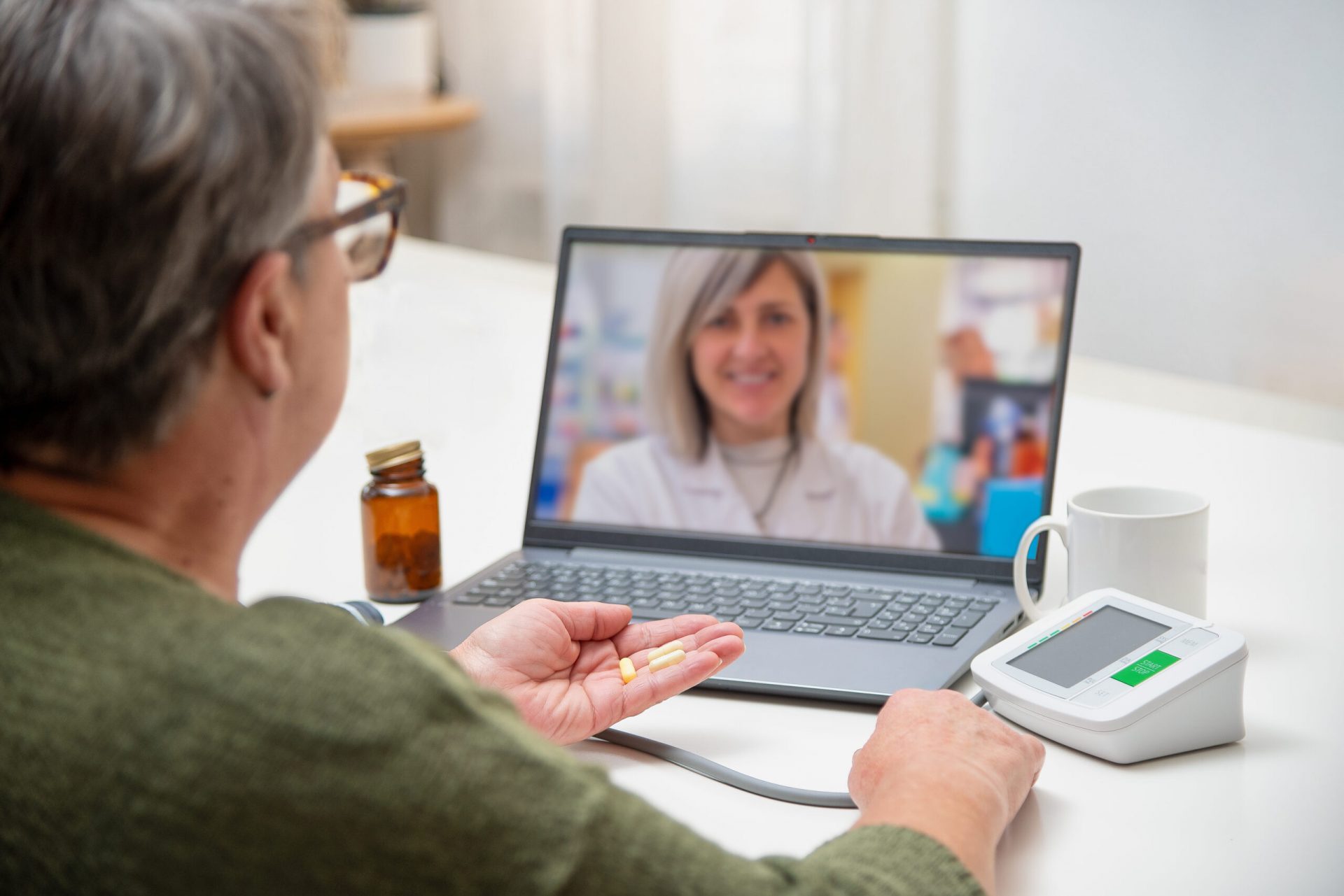 Senior woman having online medical consultation with doctor using laptop, holding pills in hand and checking blood pressure at home
